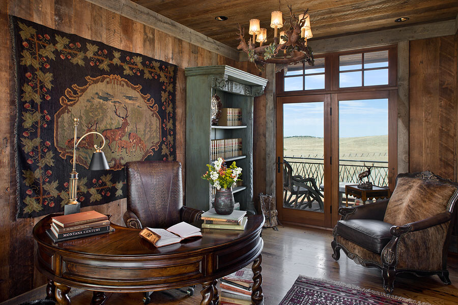 Home office with semi-circular wood desk and leather office chair. A tapestry with a deer family hangs on the wall next to a green book shelf with leather bound books. A glass door opens to a patio that overlooks rolling hills. A cow hide and leather armchair sits across from the desk.