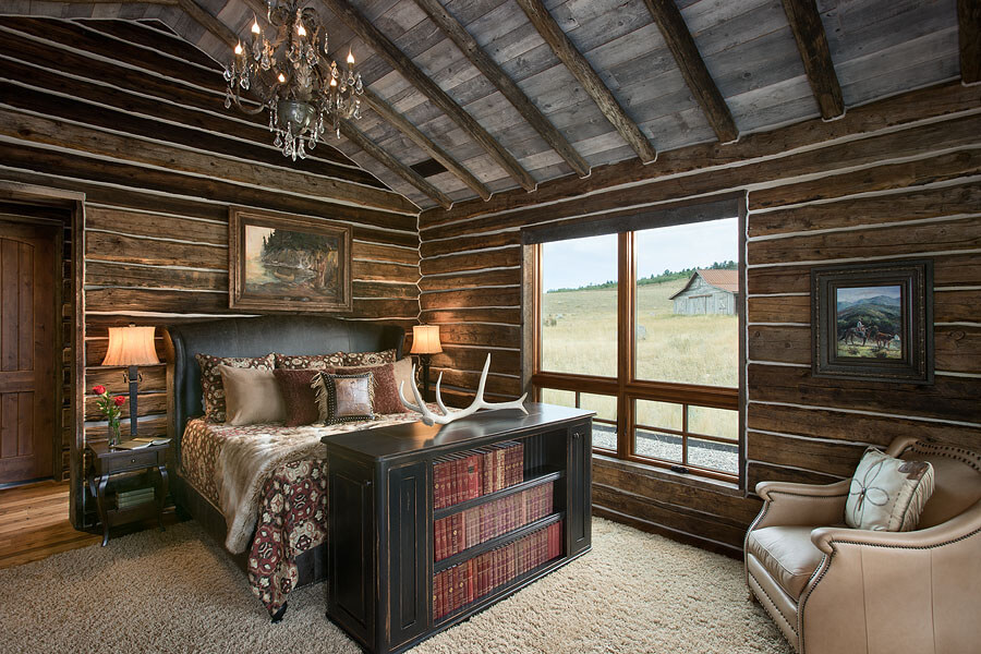 Reconstructed wood cabin as master bedroom. Dark leather bed frame with brown and neutral bedding and pillows. A low bookshelf sits at the foot of the bed with leather bound books. A light brown armchair with pillow sits in the corner under Western-style painting..