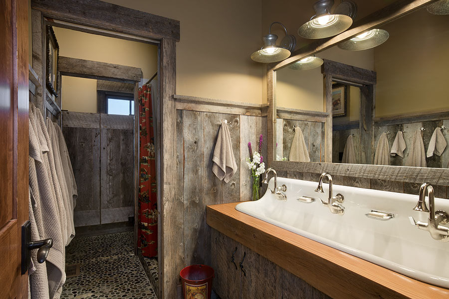 Guest bathroom with extra long basin sink below long rectangular mirror. A small standing shower is covered with red fabric curtain. Saloon doors separate shower from rest of bathroom. Gray towels hang on the wall of reclaimed wood wainscotting.