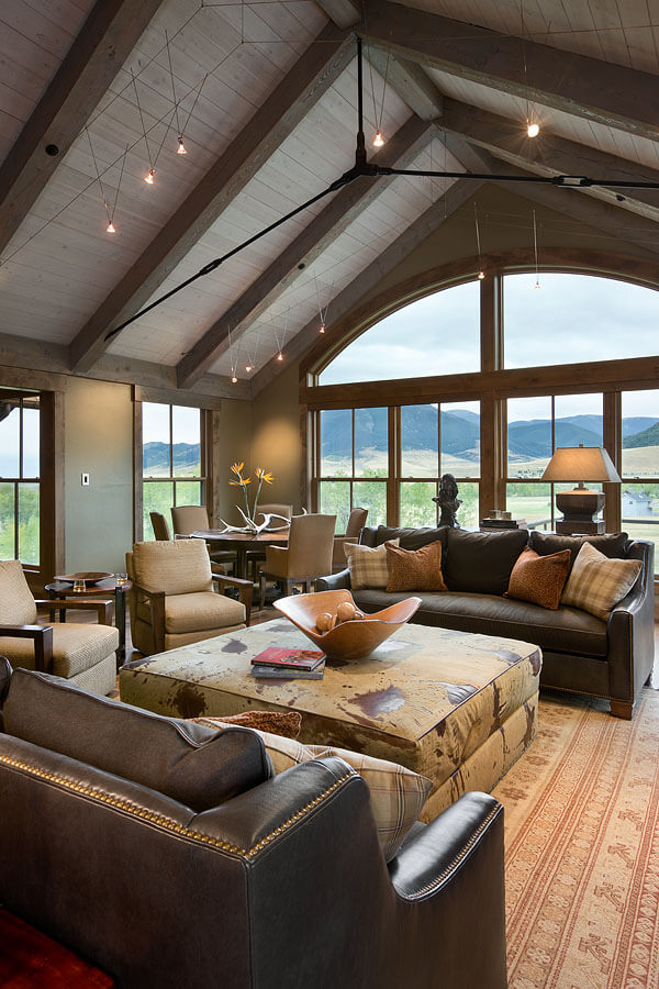Large open living room with brown leather sofas, patterned ottoman, and two armchairs. Windows face rolling hills and mountains.