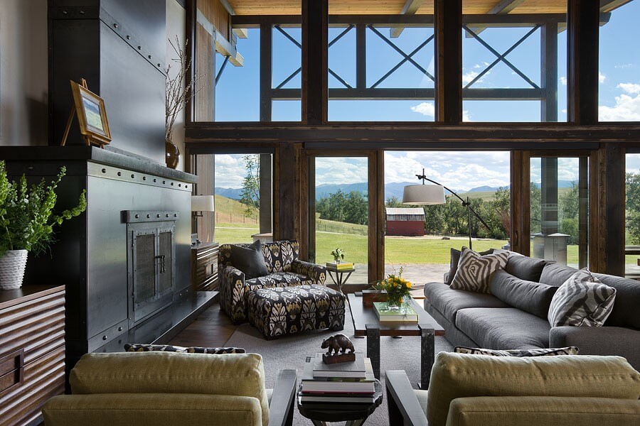 Living room with large metal fireplace, soft gray sofa, patterned armchair, coffee table, and view of barn and mountains.